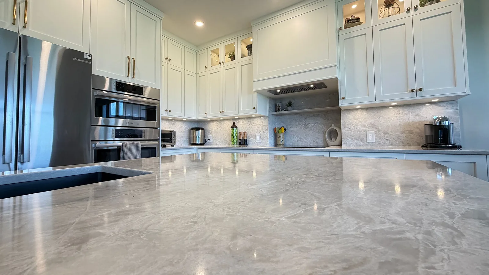 Recently completed Kelowna kitchen renovation with a quartz island, painted shaker cabinetry, lit glass uppers, and a marble-look backsplash by Amend Developments