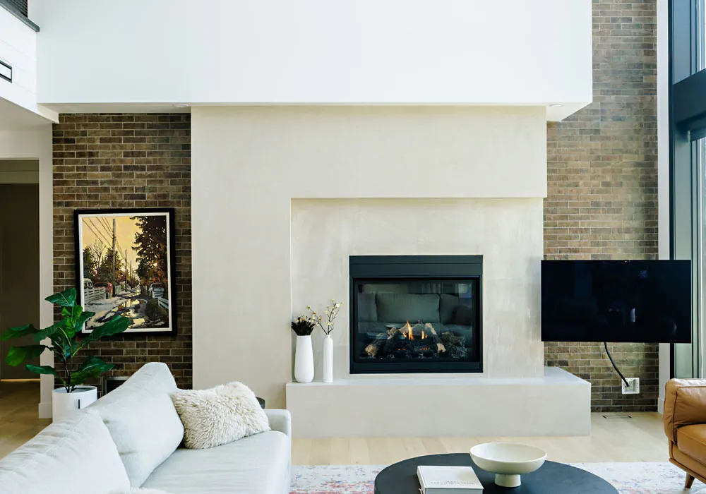 Double-height living room with a plaster fireplace surround and brick feature wall by Amend Developments
